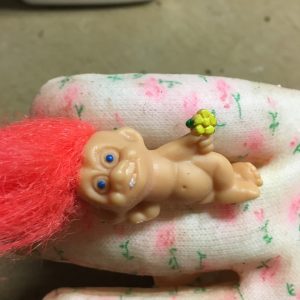 Small plastic troll with orange hair in a lying down position between two fingers of a glove.